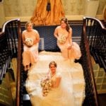 175-kapustasm Happy Bride sitting on the stairs and smiling to the photo with two bridesmaids in pastel pink dresses.