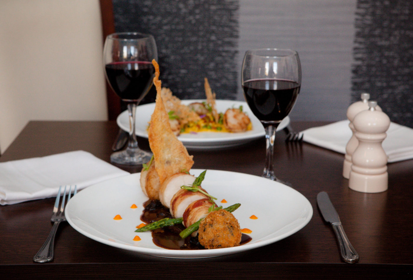 Two dinner plates set out with a knife, fork, napkin and two glasses of red wine.